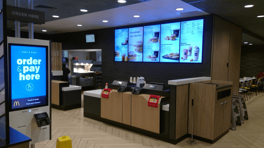 Indoor image of McDonalds showing an order kiosk and main menu screens.